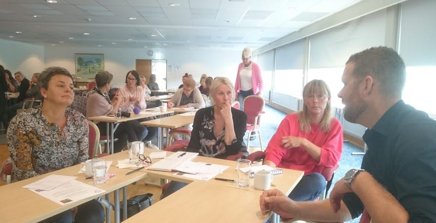 Människor som sitter i en utbildningssal kring flera mindre bord och samtalar