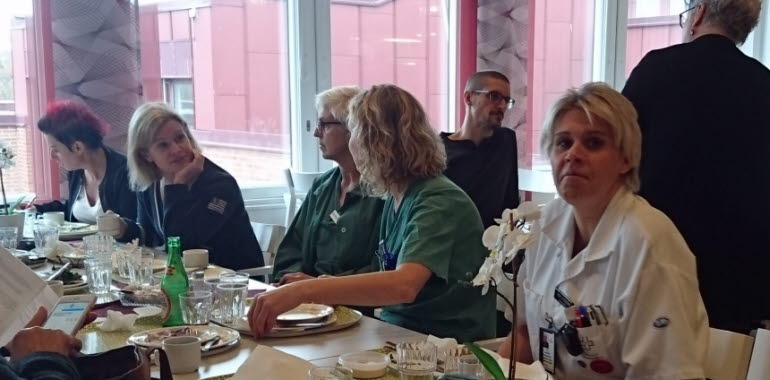 En grupp kvinnor i sjukhuskläder sitter vid ett bord och samtalar och äter