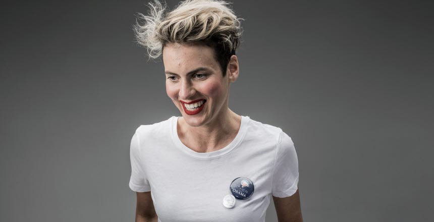 Halvkroppsbild på leende, blond kortklippt kvinna med röda läppar. Klädd i vit t-shirt. Grå bakgrund.