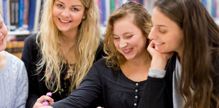 Tre unga kvinnor (studenter) sitter civilklädda och pratar och ler, en tittar in i kameran