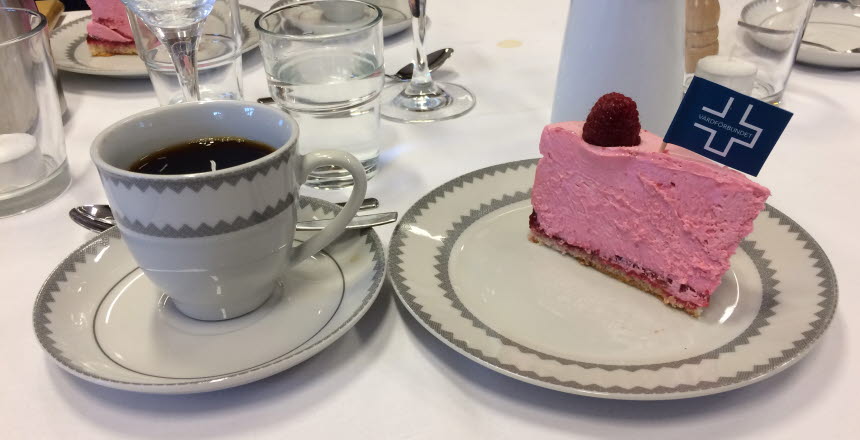En kaffekopp med kaffe i samt en tallrik med en bit tårta med hallon och en liten flagga i med Vårdförbundets logga
