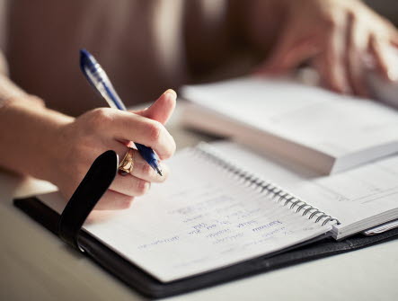 Två uppslagna anteckningsböcker på ett bord. Till vänster ser man en hand som håller i en penna.