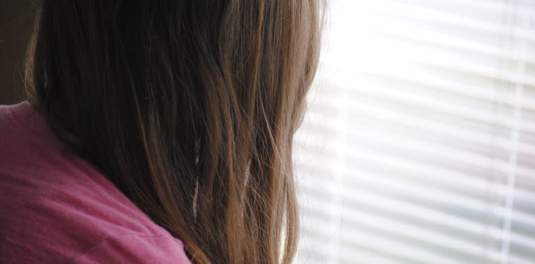 Kvinna eller flicka tittar ut genom ett fönster med neddragna persienner. Långt brunt hår döljer ansiktet.