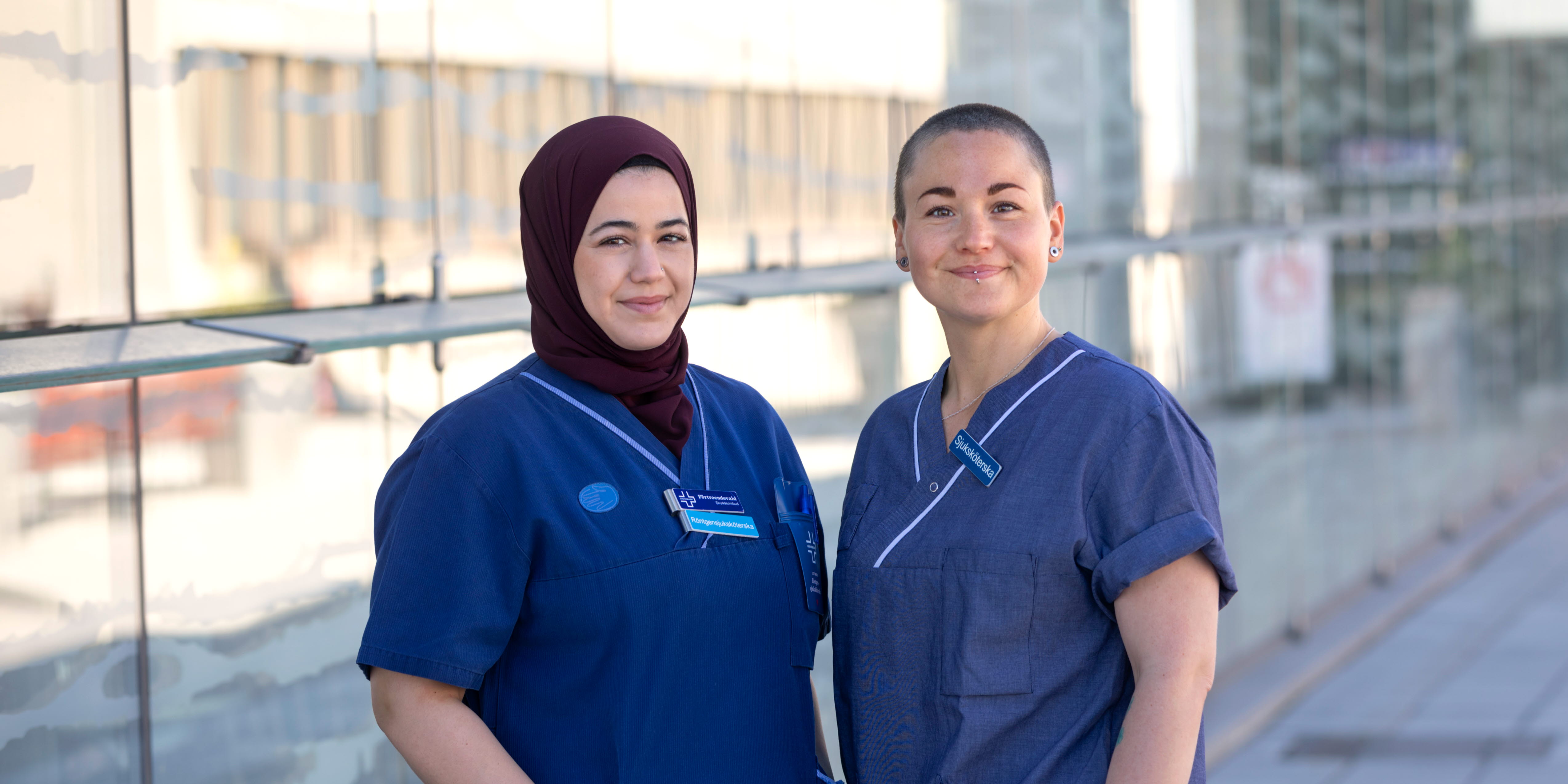 Två röntgensjuksköterskor i blå arbetskläder står utanför ett sjukhus. De ser glada ut och tittar in i kameran.