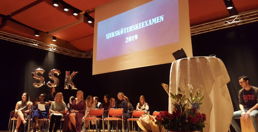 Nyutexaminerade sjuksköterskor som sitter på stolar på en scen. I bakgrunden finns det ballonger med bokstäverna SSK och på en skärm står det sjuksköterskeexamen 2019