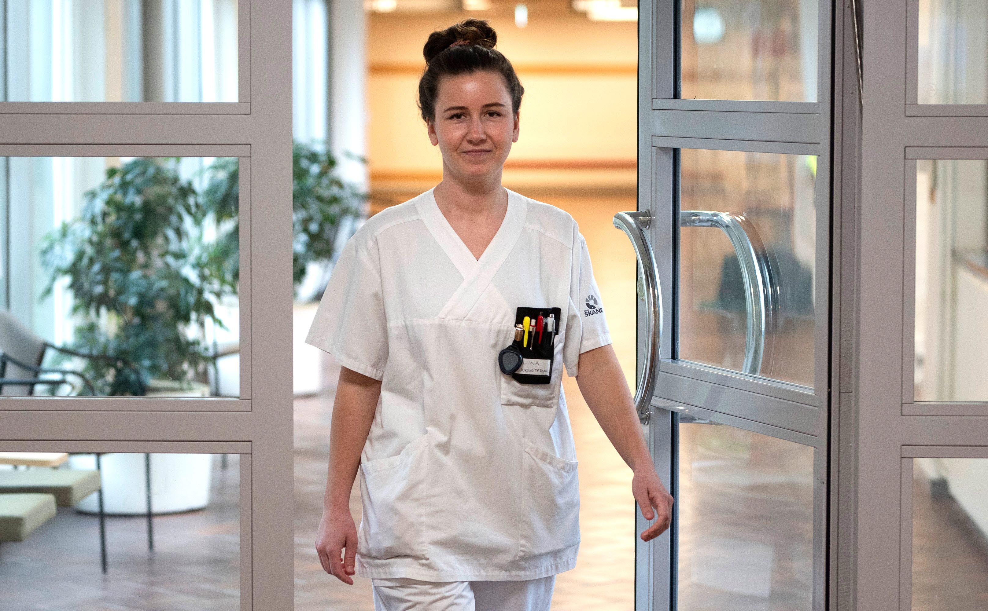 Sjuksköterska passerar genom dörr . Hon har mörkt hår i en knut på huvudet och vita arbetskläder.